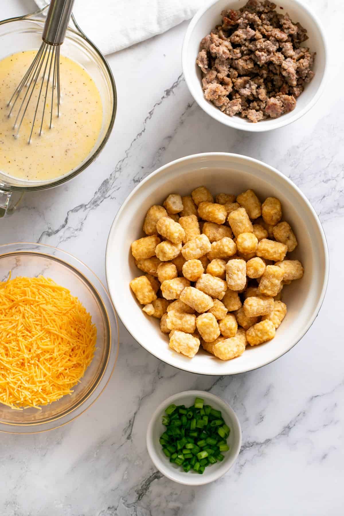 egg mixture, tater tots in a bowl, crumbled sausage, shredded cheese and green onion.